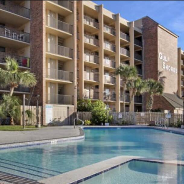 A modern multi-story hotel named Gulf Shores stands behind a clear outdoor swimming pool, surrounded by palm trees, balconies, and a sunlit patio area.