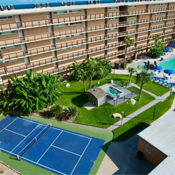 Aerial view of a hotel courtyard featuring a blue tennis court, a hot tub, palm trees, grassy areas, lounge chairs, and a large swimming pool with blue umbrellas. Multiple floors of hotel rooms surround the courtyard.