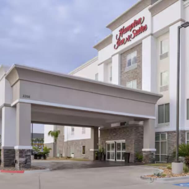 Exterior view of a Hampton Inn & Suites hotel with a covered entrance, large windows, stone accents, and a well-maintained driveway with some landscaping and palm trees.