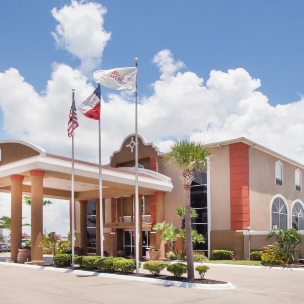A tan and white hotel with arched windows, tall columns at the entrance, three flagpoles flying flags, palm trees, a fountain, and a clear blue sky with scattered clouds.