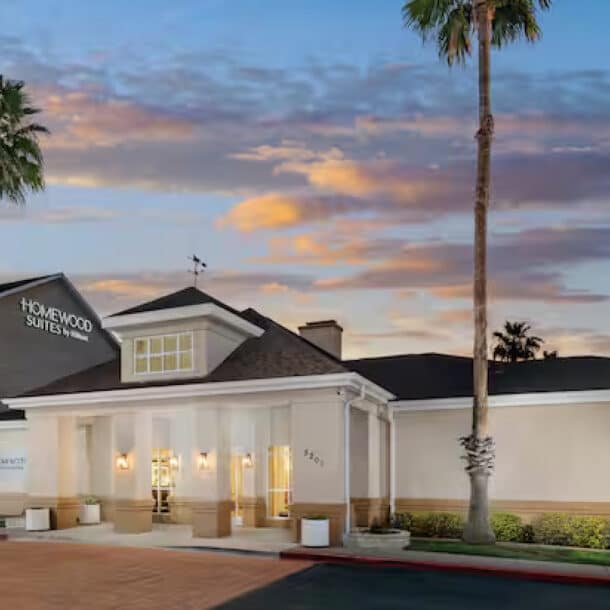 A Homewood Suites hotel building at sunset with palm trees, an American flag, and a well-lit entrance. The sky is colorful with clouds, and the hotel signs are visible on the building and a flagpole.