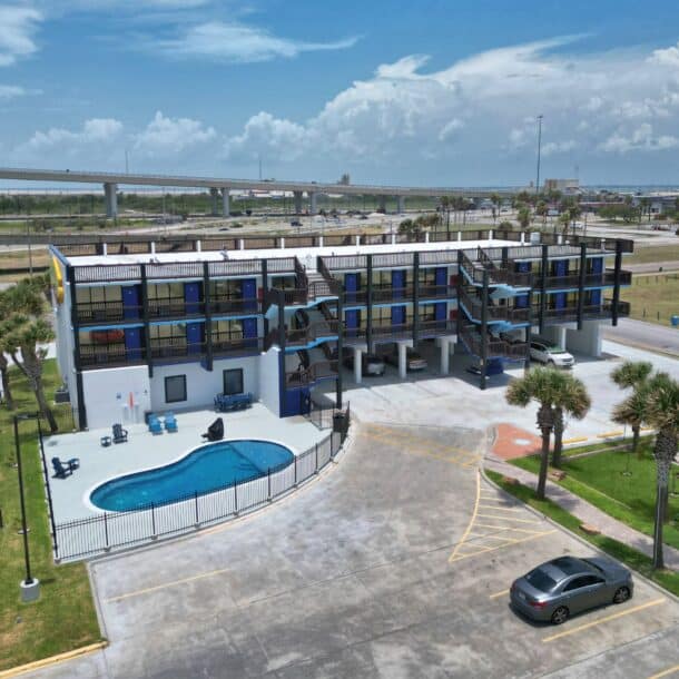 Aerial view of a modern three-story motel with blue balconies, a fenced outdoor swimming pool, several parked cars, and palm trees, set near a highway overpass under a partly cloudy sky.