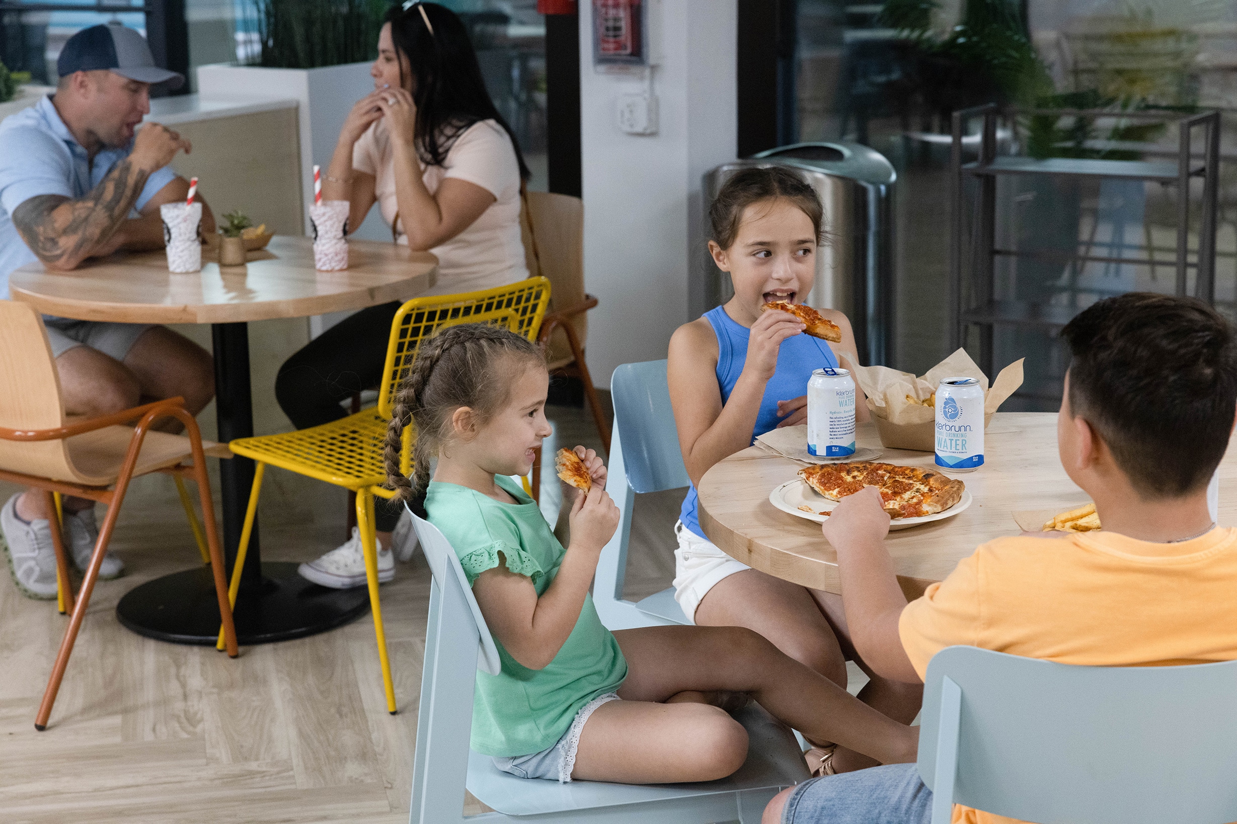Three children sit at a table eating pizza and drinking water, while a man and woman talk at another table in the background. The scene appears to be inside a casual dining restaurant or café.