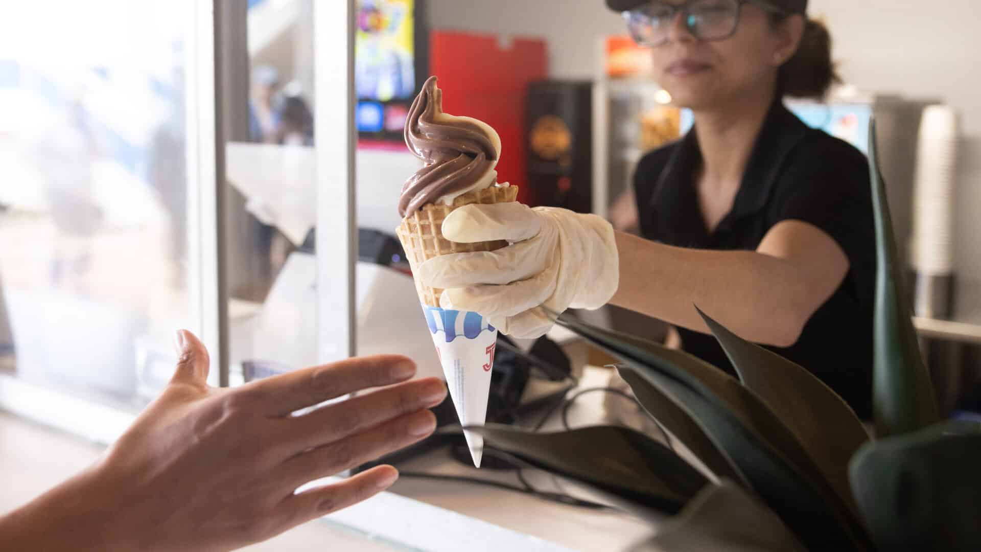 A person wearing gloves hands a chocolate-vanilla swirl ice cream cone in a waffle cone to a customer reaching out over a counter at an ice cream shop.