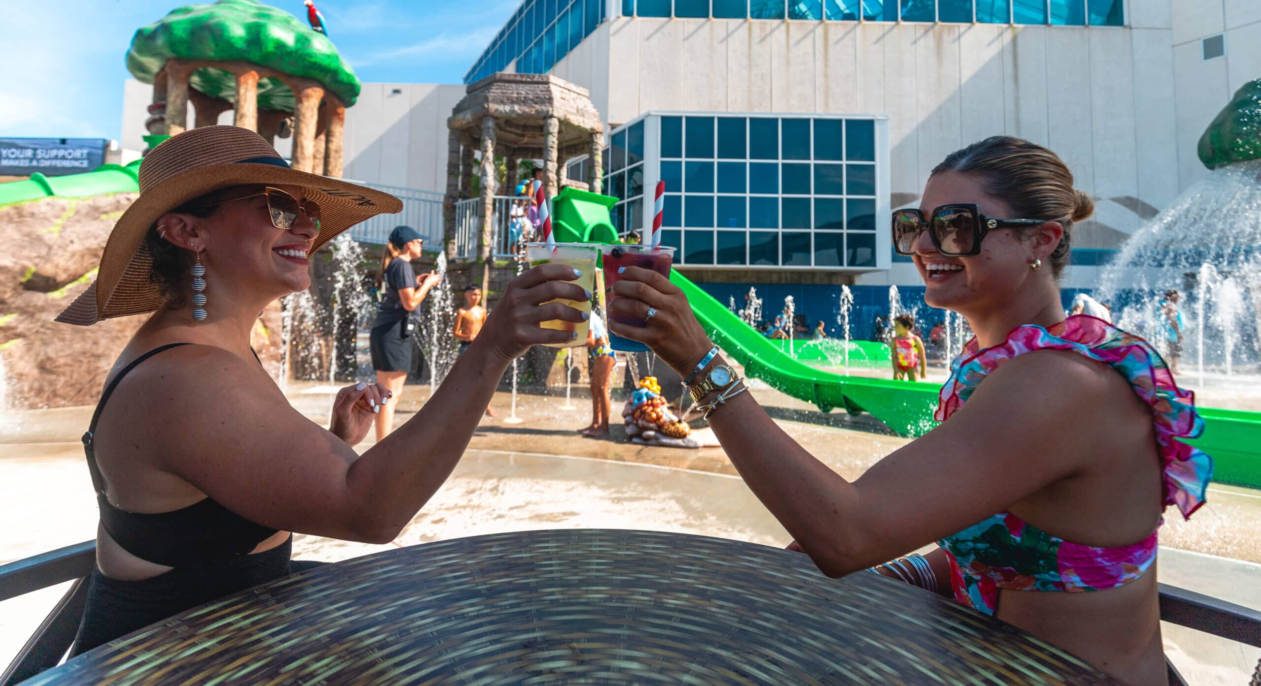 Two women in swimsuits and sunglasses smile and toast with drinks at an outdoor table near water slides and fountains at a water park on a sunny day.