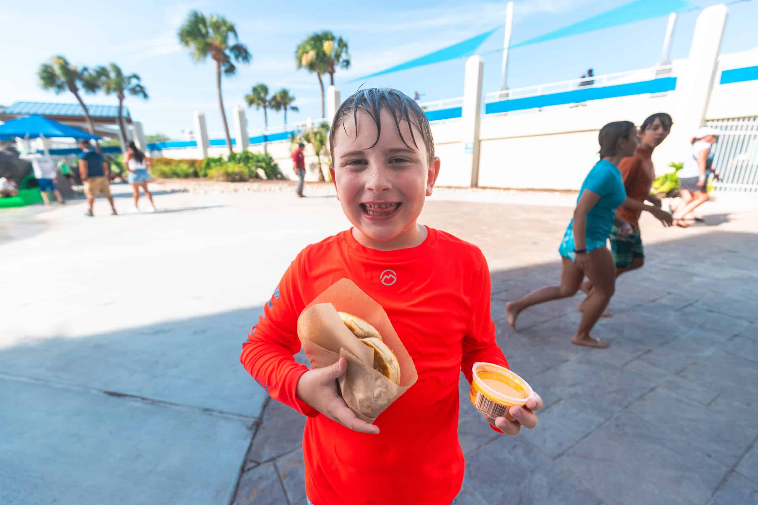 A smiling child in a bright orange rash guard holds a sandwich and a cup of cheese dip at an outdoor water park, with palm trees and people in swimwear in the background.