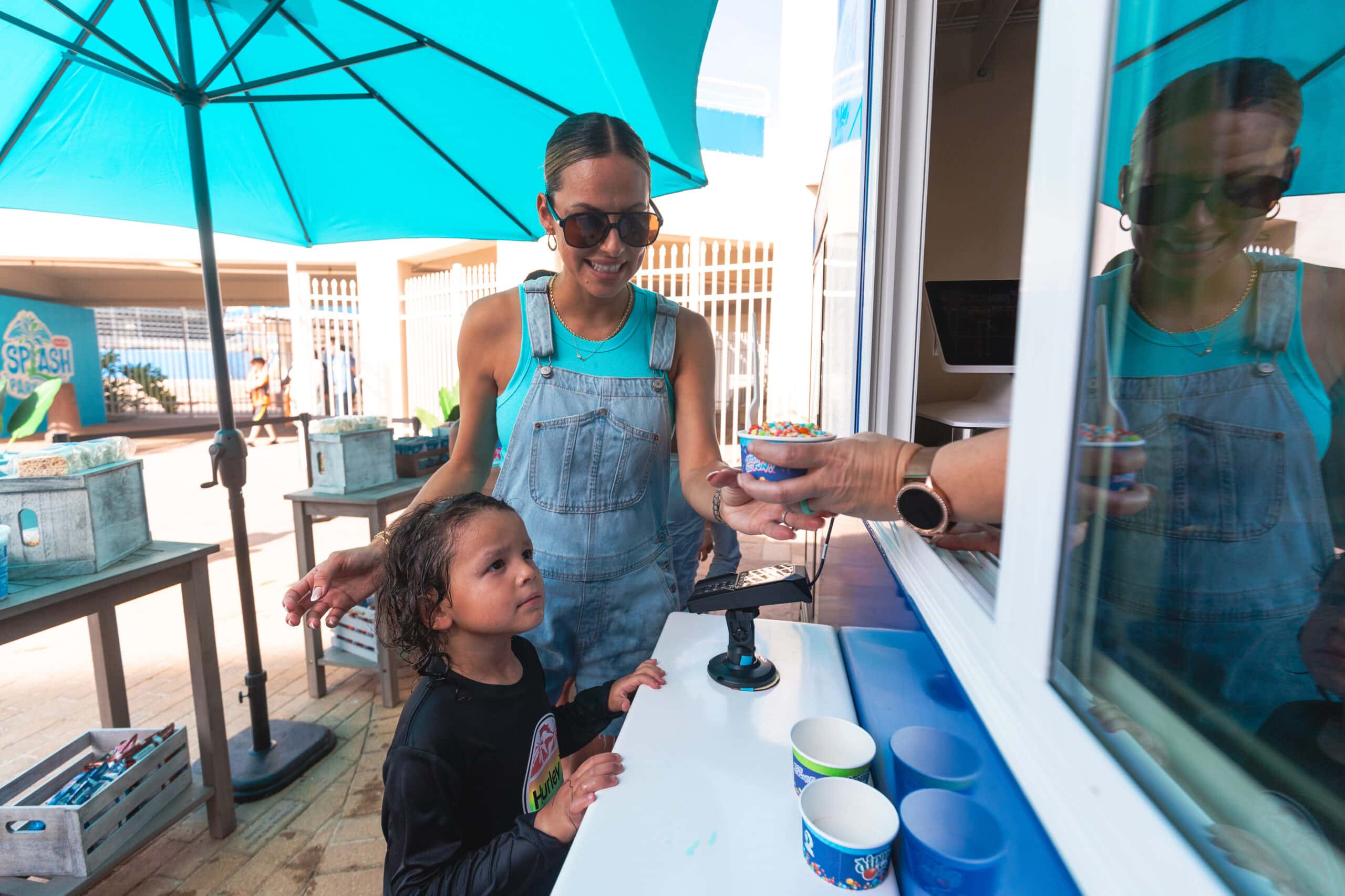A woman and a young child stand at an outdoor service window. The woman takes a bowl of ice cream from a person inside while the child looks up, waiting. Blue umbrellas and tables are in the background.