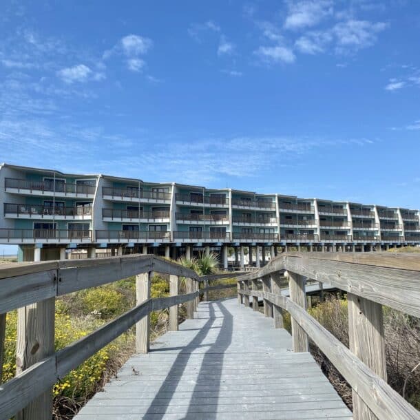 A wooden boardwalk leads through dunes and yellow wildflowers toward a modern, multistory beachfront building elevated on stilts, under a bright blue, partly cloudy sky.