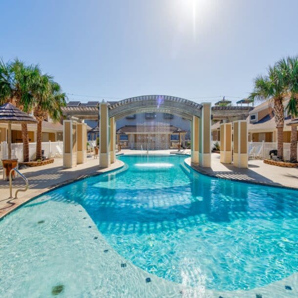 Resort-style outdoor swimming pool with clear blue water, palm trees, lounge chairs, and a covered bridge feature, surrounded by modern two-story buildings under a bright sunny sky.