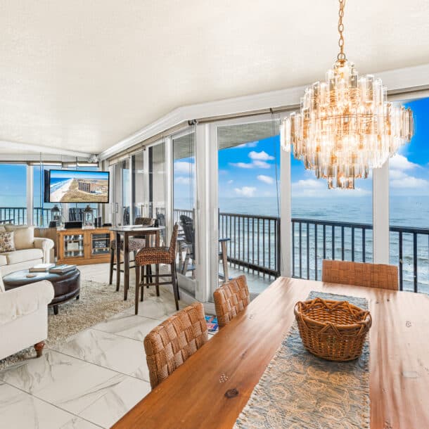 A bright beachfront living and dining area with large windows offering ocean views, a chandelier over a wooden table, wicker chairs, a cozy sofa, and a telescope facing the water.
