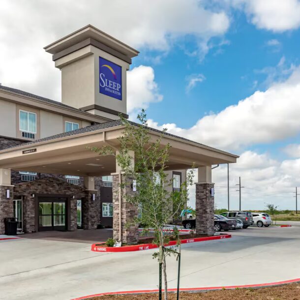 A modern Sleep Inn hotel building with a covered entrance, stone accents, and a parking lot with several cars under a partly cloudy sky.