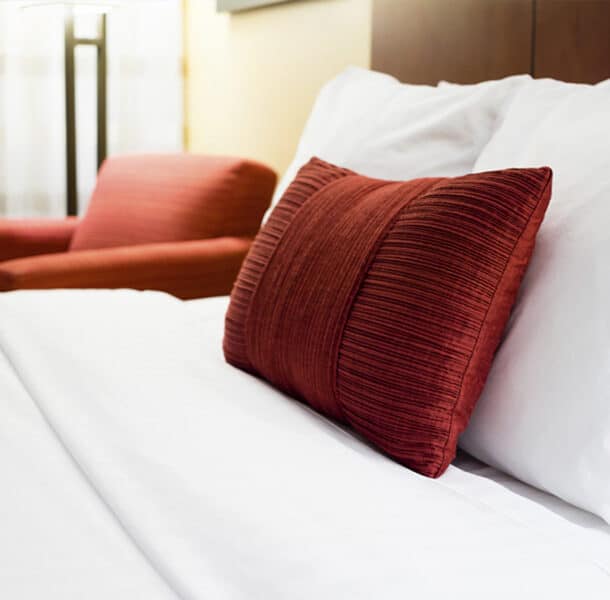 A neatly made bed with white sheets and pillows, featuring a textured red accent pillow. In the background, there is a matching red armchair and a softly lit room.