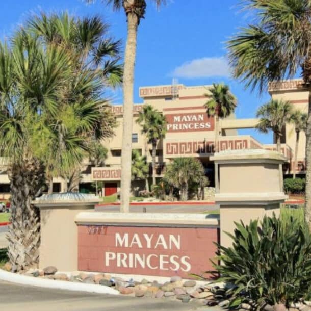 Entrance to the Mayan Princess resort, featuring a sign with red lettering, surrounded by palm trees, plants, and a multi-story tan building with decorative details in the background.