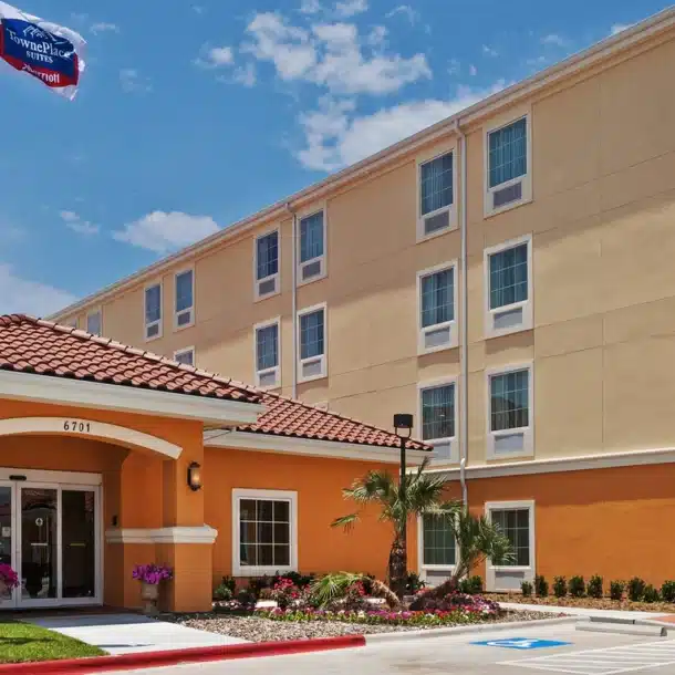 A four-story TownePlace Suites by Marriott hotel with a yellow and orange exterior, three flagpoles, palm trees, and a parking lot with accessible parking spaces in front, under a blue sky with clouds.