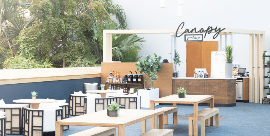 A bright, modern café area with wooden tables, chairs, potted plants, and a counter labeled Canopy pickup. Bottles and coffee machines are visible behind the counter, and large leafy plants surround the space.