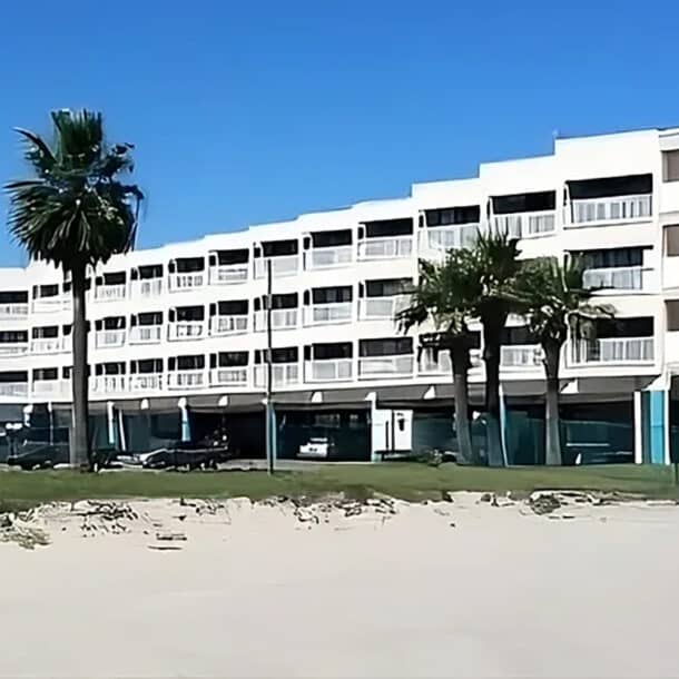 A large, white multi-story building with balconies stands behind palm trees and a sandy area under a clear blue sky. Cars are parked in front of the building.