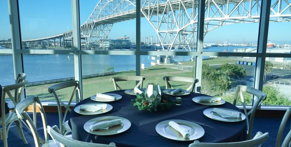 A round dining table set for six with white plates and napkins sits by large windows overlooking a river and a large arched bridge on a sunny day.