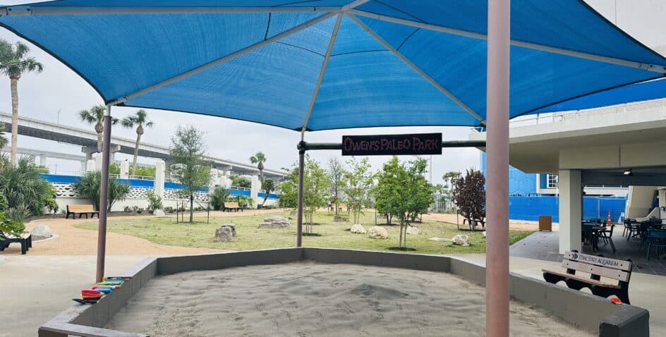 A covered sandbox labeled Owens Paleo Park sits in an outdoor area with trees, benches, and a nearby building. A blue canopy provides shade, and several toy buckets are lined up along the sandboxs edge.