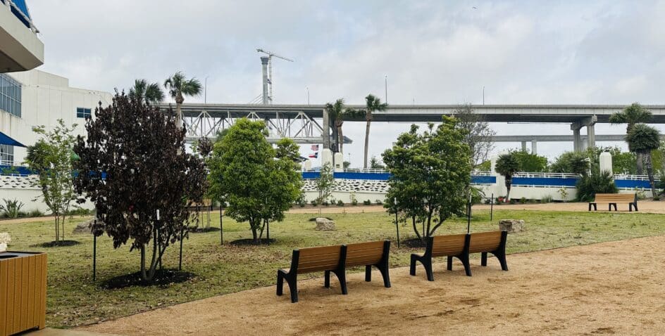 A small park area with two wooden benches, several green and brown trees, a sandy path, and a large elevated highway in the background under a cloudy sky.