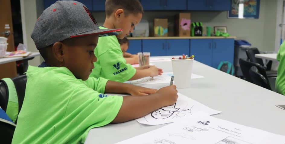 Young children wearing green shirts sit at a table coloring pictures with crayons in a classroom. Art supplies and colorful drawings are visible on the table and in the background.