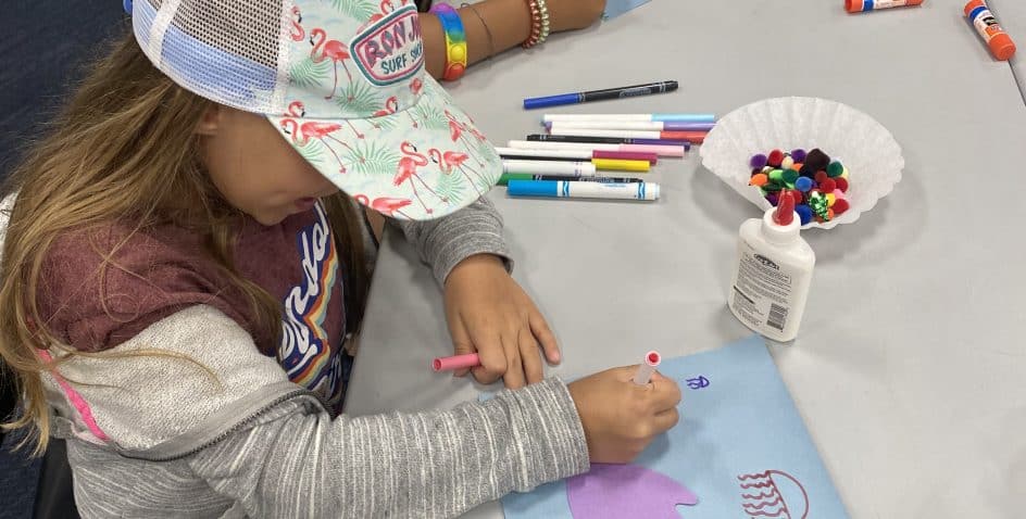 A young child wearing a flamingo cap colors on blue paper at a table with markers, glue, pom-poms, and craft supplies. Another child’s hands with bracelets are visible nearby.