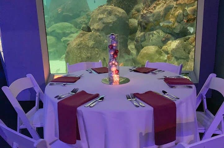 A round table with a white tablecloth is set for five, each place with red napkins and silverware. A tall centerpiece with ornaments is in the middle. Large aquarium windows with rocks and fish are in the background.