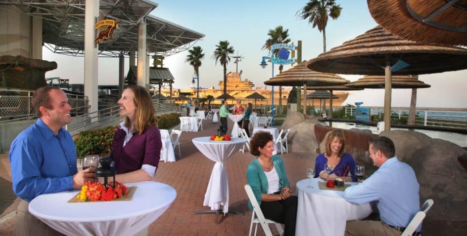People enjoy food and drinks at outdoor tables near the ocean, with palm trees, umbrellas, and a boardwalk in the background. The atmosphere is lively and relaxed, with a clear sky and warm lighting.