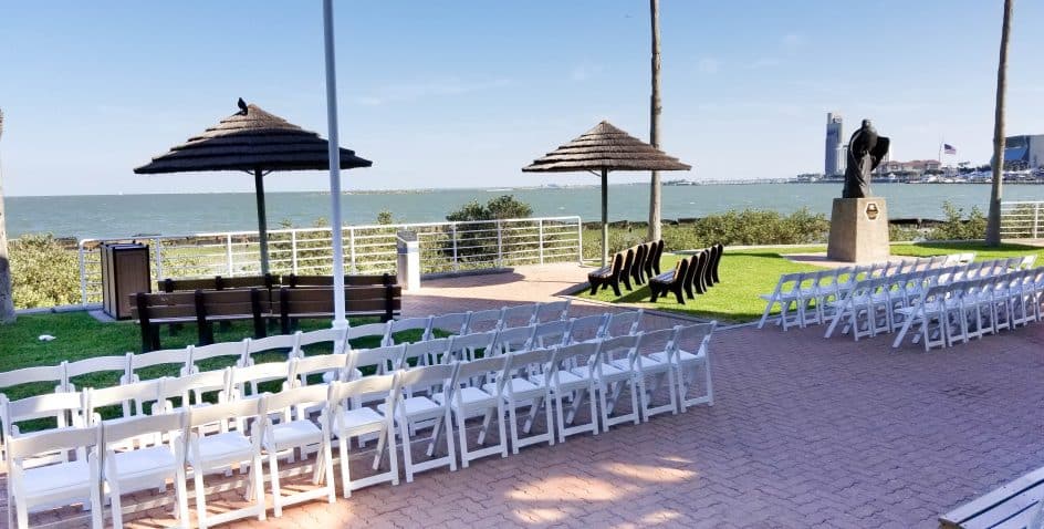 Rows of white chairs are set up outdoors facing a statue near the waterfront, with the ocean, umbrellas, and distant city buildings visible under a clear blue sky.
