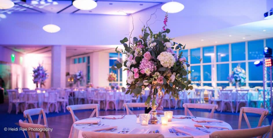Elegant wedding reception hall with round tables, white chairs, and large floral centerpieces. Soft purple and blue lighting creates a romantic ambiance, and candles add a warm glow to the table setting.