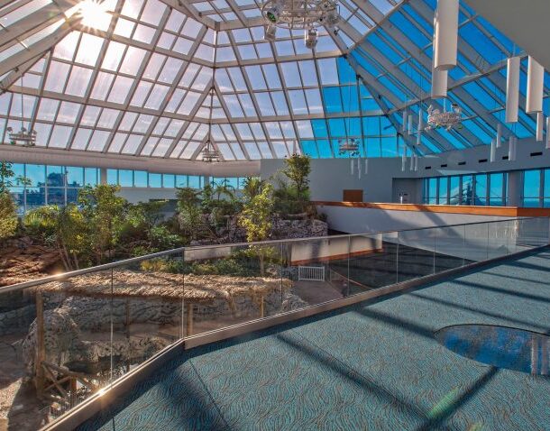 A spacious indoor atrium with a glass ceiling, filled with sunlight, lush plants, trees, and a rocky landscape. There’s a blue-carpeted walkway with a glass railing, creating a bright, inviting atmosphere.