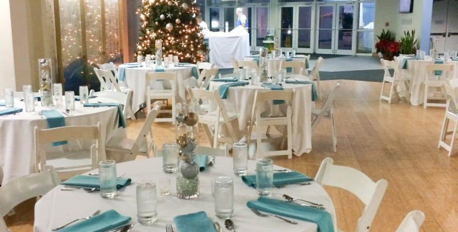 A banquet hall decorated for an event, featuring round tables with white tablecloths, turquoise napkins, glassware, and festive centerpieces. A decorated Christmas tree is in the background near large windows.