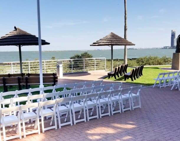 Rows of white chairs are set up outdoors facing a waterfront, with a statue on a pedestal, two thatched shade structures, and a city skyline visible in the distance under a clear blue sky.