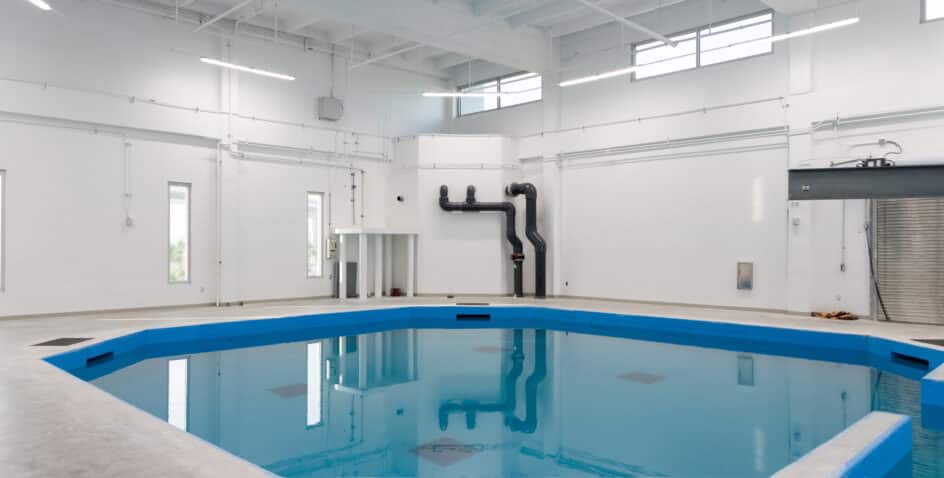 A large indoor facility with white walls and exposed ceiling pipes features a blue-edged hexagonal pool of still water in the center, surrounded by bright lighting and industrial equipment.