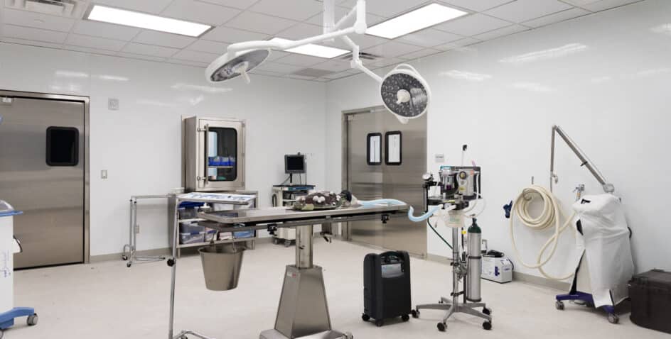 A clean, modern operating room with an empty surgical table in the center, overhead surgical lights, medical equipment, storage cabinets, and double doors in a brightly lit, sterile environment.