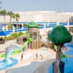 Children play in a water-themed splash pad area with dolphin and tree sculptures, slides, and fountains, surrounded by tables with blue umbrellas and palm trees. A large building and ship are visible in the background.