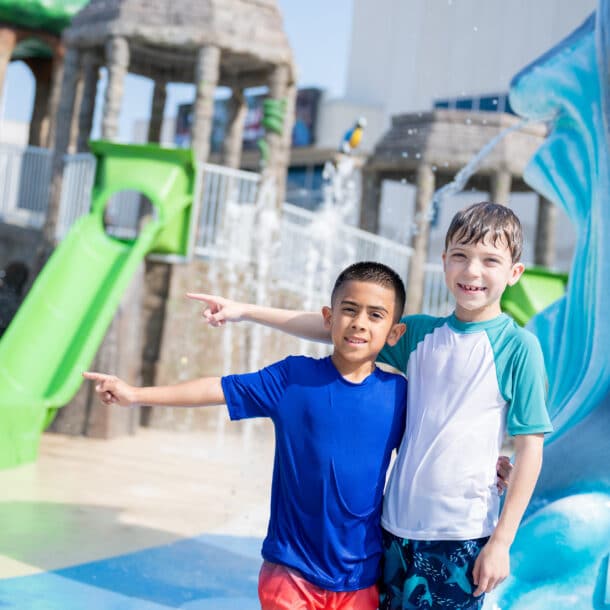 Two boys in swimsuits stand together, smiling and pointing at something off-camera at a water park. There are slides, splash features, and playful structures in the background.