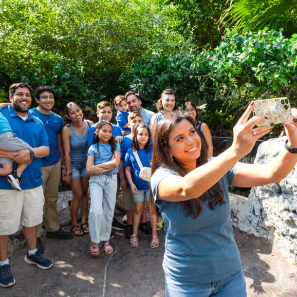 A woman takes a selfie with a group of 15 people, including children and adults, all smiling outdoors among greenery and rocks, dressed mostly in blue and casual attire.