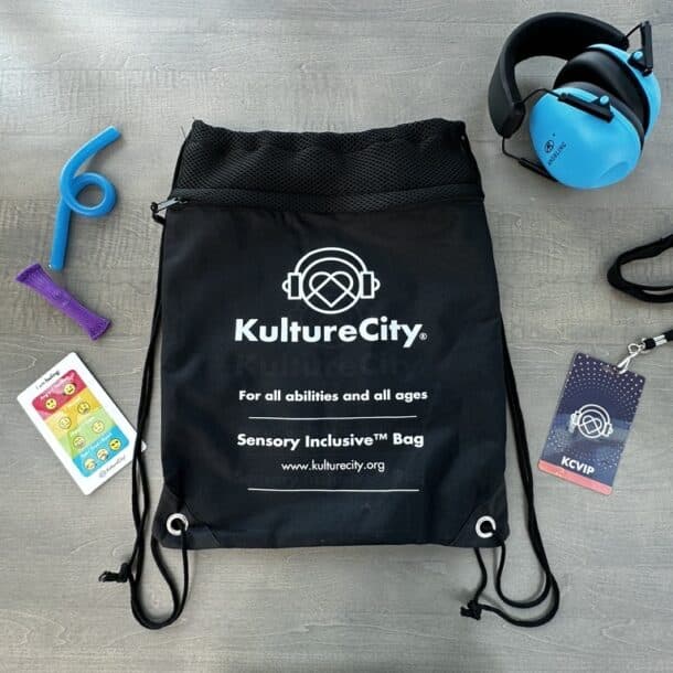 A black drawstring bag labeled KultureCity Sensory Inclusive™ Bag lies on a gray surface, surrounded by blue noise-canceling headphones, a chewable tool, a fidget toy, a sensory card, and a KCVIP badge on a lanyard.
