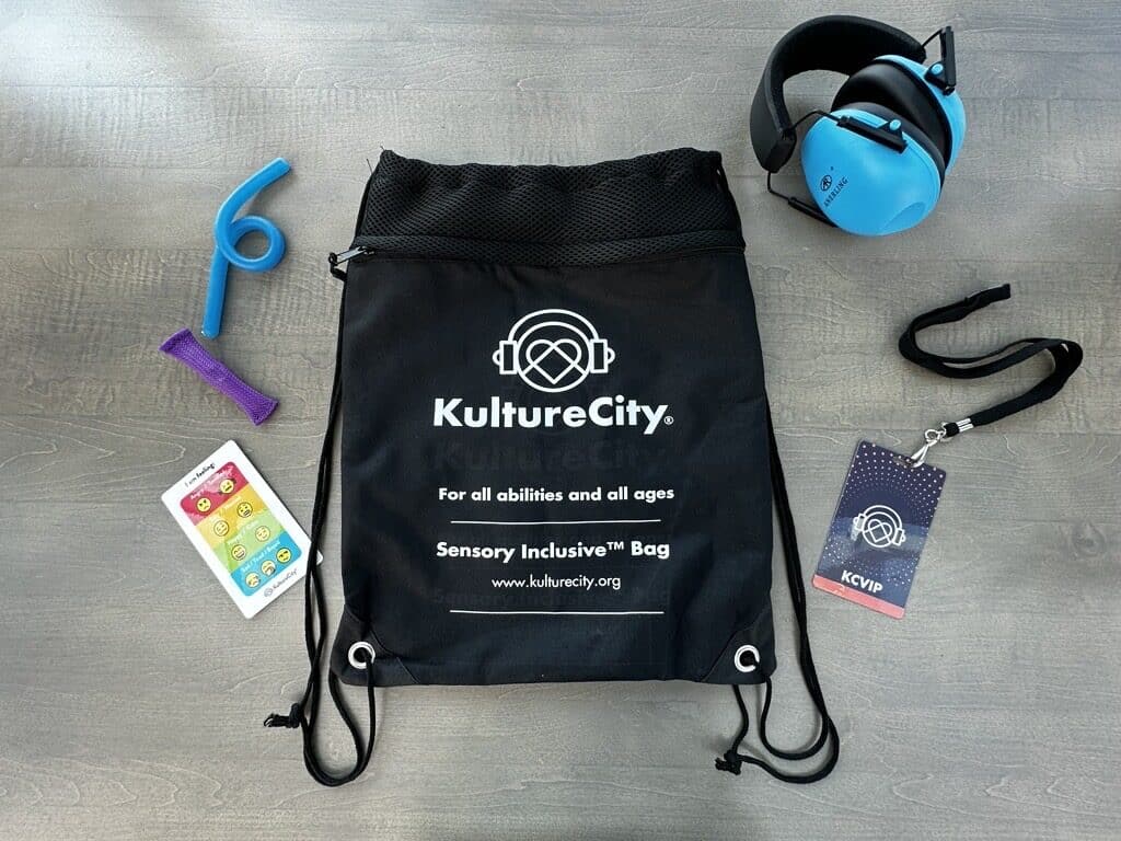 A black drawstring bag labeled KultureCity Sensory Inclusive™ Bag lies on a gray surface, surrounded by blue noise-canceling headphones, a chewable tool, a fidget toy, a sensory card, and a KCVIP badge on a lanyard.