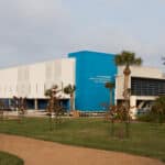 A modern building with white and blue walls labeled “Texas State Aquarium Center for Wildlife Rescue” stands behind a grassy area with young trees and a dirt pathway under a cloudy sky.