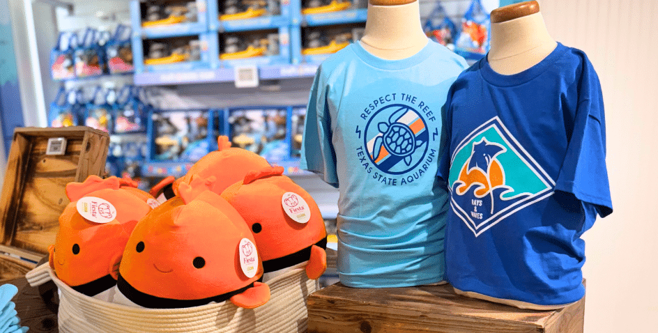 Two mannequins display blue t-shirts with aquatic-themed prints, next to a basket filled with orange plush fish toys in a gift shop with marine-themed merchandise.