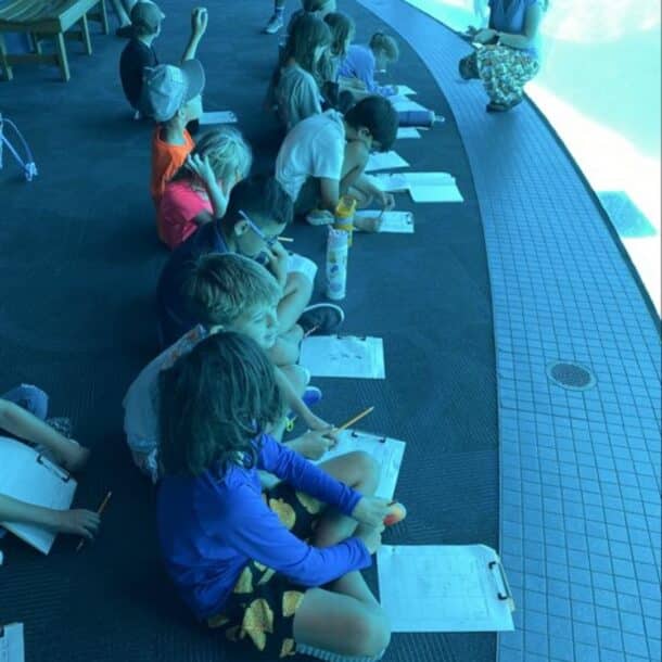 A group of children sit on the floor with clipboards and pencils, facing a woman near a large aquarium window. They appear to be observing and taking notes, possibly for an educational activity.