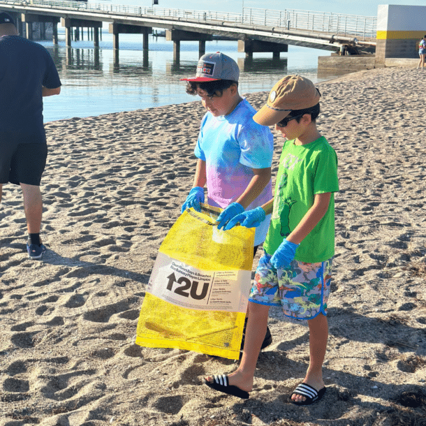 Two boys wearing gloves collect trash in a yellow bag on a sandy beach. They walk near the shoreline with a pier and other people visible in the background on a sunny day.