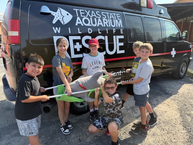 Six children smile and pose in front of a Texas State Aquarium Wildlife Rescue van, holding a plush shark and a green net, suggesting a fun, educational activity related to animal rescue.