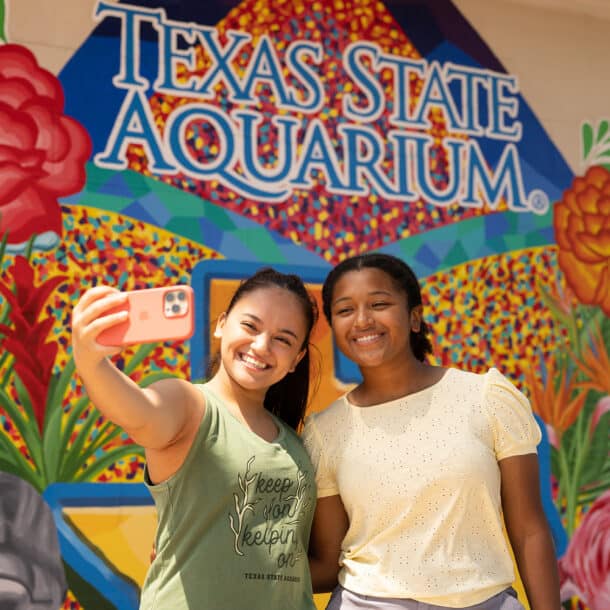 Two smiling young women take a selfie in front of a colorful mural that reads Texas State Aquarium, featuring flowers, an elephant, and a flamingo.