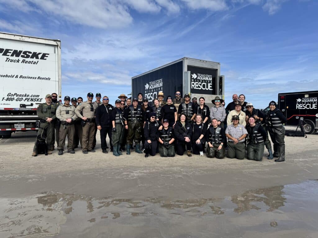 A large group of uniformed people and a dog pose together on a beach in front of trucks labeled Penske and Texas Parks and Wildlife Rescue under a partly cloudy sky.