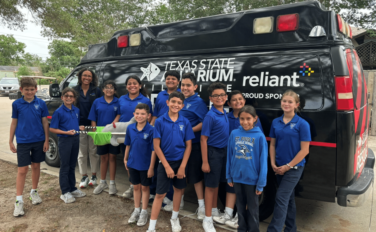 A group of students in blue shirts stand and smile in front of a black Texas State Aquarium vehicle, holding a green net, with trees and buildings in the background.