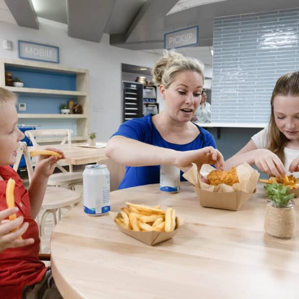 A woman and two children sit at a table eating fries and fried food from paper trays, with cans of soda. They appear to be in a bright, modern cafe or cafeteria.