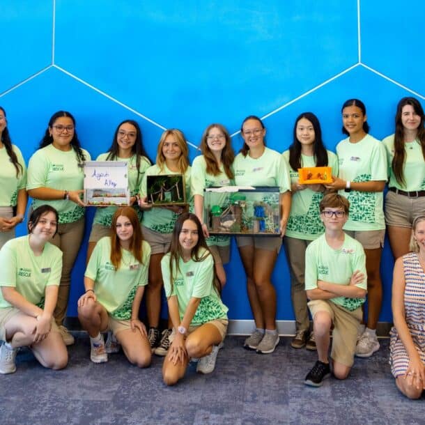 A group of 13 young people and one adult sit and stand in front of a blue wall, wearing matching light green shirts. Some hold colorful handmade dioramas. They are smiling and posing for a group photo indoors.