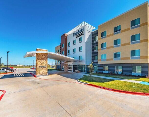 A modern Fairfield Inn & Suites hotel building with four stories, beige and white exterior, a covered entrance, landscaped grass, and a clear blue sky above. A few cars are parked nearby.